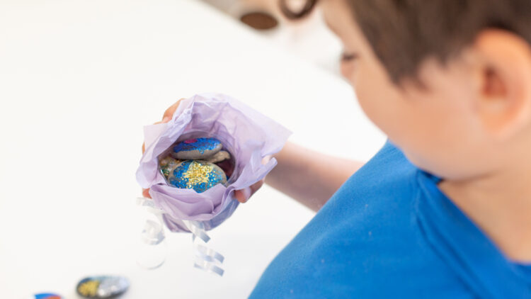 男孩拿着装有薄纸的容器，里面装满了彩绘的岩石。